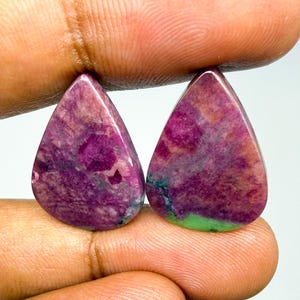 May include: Two pear-shaped cabochon gemstones featuring a mottled blend of deep magenta, purple, and dark red hues with visible inclusions. One stone displays a distinct patch of vibrant green mineral at the base, while both have a smooth, polished surface.