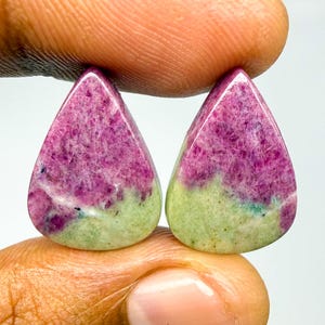 May include: Two pear-shaped cabochons featuring a distinct color split of vibrant magenta-pink and pale lime-green. These polished gemstones display a natural crystalline texture with visible mineral inclusions throughout the surface of each stone.