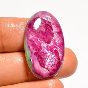 May include: An oval-shaped cabochon gemstone featuring a vibrant magenta and deep pink center with a translucent, light green border. The surface displays natural internal fractures and a polished finish with a distinct white light reflection.