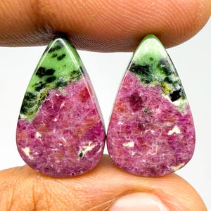 May include: Two pear-shaped ruby zoisite gemstones featuring a vibrant magenta-pink base with contrasting patches of bright green and dark black mineral inclusions. The stones have a smooth, polished surface and a distinct teardrop silhouette.