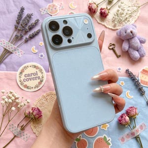 May include: A pale blue phone case with a textured back and a camera module. The case has a smooth, matte finish and multiple camera cutouts. The text "carol covers" is visible on a circular sticker. The case is designed to protect a smartphone.