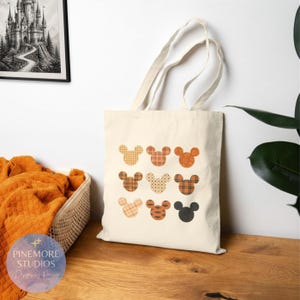 Peut inclure: Un sac fourre-tout en toile couleur crème orné d'une grille de neuf silhouettes d'oreilles de souris. Chaque silhouette présente un motif unique dans des tons orange, marron et noir, incluant des carreaux, des pois, des rayures et du noir.