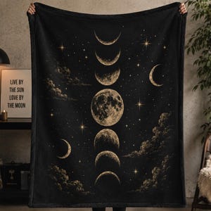 May include: A black rectangular blanket featuring a vertical sequence of moon phases in varying shades of gold and cream. The design includes scattered stars and detailed clouds against a dark background, creating a celestial-themed pattern.