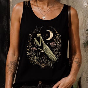 Puede incluir: Una camiseta de tirantes negra con un estampado gráfico central de una mantis religiosa verde rodeada por una disposición circular de elementos botánicos dorados, pequeñas estrellas y una luna creciente sobre tela oscura.