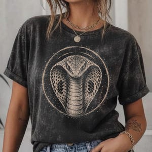 Può includere: Una t-shirt grigio antracite con effetto lavaggio acido, caratterizzata da una grafica circolare di un cobra al centro. Il capo presenta maniche corte e un girocollo. Il disegno del cobra è realizzato in uno stile puntinato dettagliato con accenti dorati.