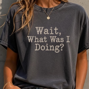 Puede incluir: Una camiseta gris carbón de manga corta con el texto blanco desgastado "Wait, What Was I Doing?" impreso en la parte delantera. La prenda tiene cuello redondo y un corte holgado, combinada con unos vaqueros azules.
