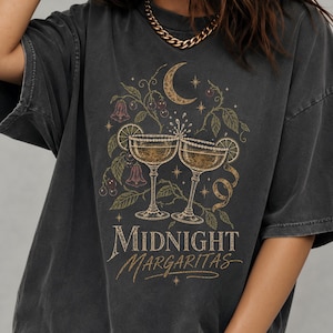 May include: A charcoal grey short-sleeve t-shirt featuring a graphic print of two margarita glasses clinking together, surrounded by vines, flowers, and a crescent moon. The text "MIDNIGHT MARGARITAS" is printed in a gold-toned font below the glasses.