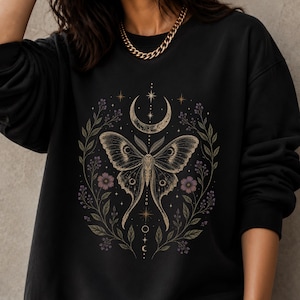 May include: A black long-sleeve sweatshirt featuring a central graphic print of a moth surrounded by a crescent moon, stars, and botanical elements including leaves and small purple flowers in a circular arrangement with fine line details.
