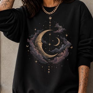May include: A black crewneck sweatshirt featuring a central graphic of a crescent moon with a human face, surrounded by dark purple and grey clouds, small stars, and a vertical alignment of moon phases extending above and below the main design.