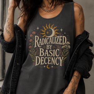 Könnte beinhalten: Ein ärmelloses Tanktop in Anthrazit mit einem Grafikdruck, der den Text "RADICALIZED BY BASIC DECENCY" in einer Vintage-Schrift zeigt. Das Design umfasst eine Sonnen- und Mondillustration, umgeben von bunten Wildblumen und kleinen Sternen.