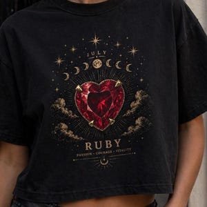 May include: Black cropped t-shirt featuring a heart-shaped ruby design with the text "JULY RUBY" and the words "PASSION COURAGE VITALITY". The shirt is worn with black jeans and a black belt.