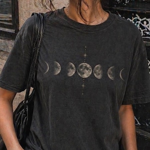 May include: Dark grey t-shirt with a moon phases graphic. The outfit includes black jeans and sunglasses. The word "Coffee" is visible on a window in the background.