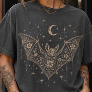 May include: Dark grey t-shirt with a bat design, floral details, a crescent moon, and stars. The bat's wings are decorated with flowers. The shirt has a vintage, worn appearance.