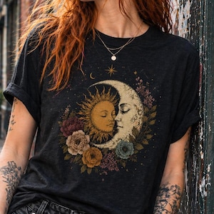Könnte beinhalten: Dunkelgraues T-Shirt mit einem Sonnen- und Mond-Design, umgeben von Blumen. Die Grafik zeigt Sonne und Mond, die sich umarmen, umgeben von Rosen und anderen Blüten. Das Shirt wird mit schwarzer Jeans und Sonnenbrille getragen.
