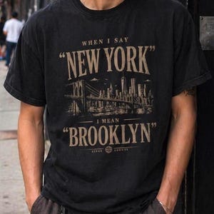 Pode incluir: Camiseta preta com o texto "WHEN I SAY NEW YORK I MEAN BROOKLYN" em estilo vintage. O design inclui uma ponte e o horizonte da cidade. A camiseta é usada com calças marrons e chaves.