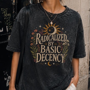 以下が含まれることがあります： 「RADICALIZED BY BASIC DECENCY」という文字の周りに太陽、月、花のグラフィックがプリントされた、チャコールグレーのアシッドウォッシュ加工Tシャツ。クルーネック、半袖、ゆったりとしたリラックス感のあるシルエットが特徴です。