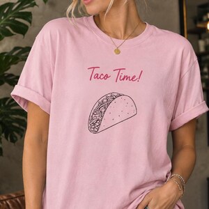 May include: A light pink short-sleeved t-shirt featuring the text "Taco Time!" in a dark pink script font above a black line-art illustration of a taco. The shirt has a crew neckline and rolled sleeves, paired with distressed blue denim jeans.