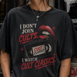 May include: Black t-shirt with the text "I DON'T JOIN CULTS. I WATCH CULT CLASSICS." A red graphic of lips and a VHS tape are also on the shirt. The person is wearing sunglasses and a silver necklace.