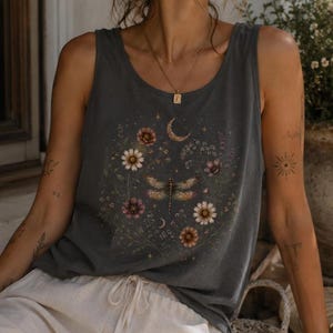 Puede incluir: Una camiseta sin mangas de color gris carbón con un estampado gráfico central de una libélula rodeada de flores silvestres, pequeñas estrellas y una luna creciente. El diseño está dispuesto en un patrón circular en la parte frontal.