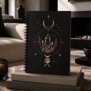 May include: A black spiral-bound notebook with a celestial design featuring a crystal cluster, crescent moons, and stars. The notebook is resting on a stack of books. The overall aesthetic is dark and mystical.
