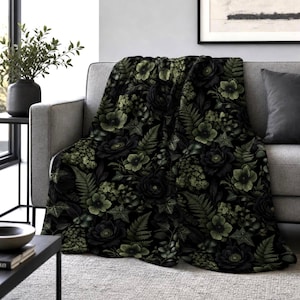 May include: A black throw blanket with a floral pattern of dark green and black roses, ferns, and other foliage. The blanket is draped over a gray sofa, creating a cozy and decorative accent for the living room.
