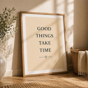 Könnte beinhalten: Ein gerahmter Kunstdruck mit hellbeigem Hintergrund und dem schwarzen Text GOOD THINGS TAKE TIME in einer Serifenschrift, ergänzt durch ein kleines Herzsymbol und zwei horizontale Linien unter dem Text, eingefasst in einen Holzrahmen.