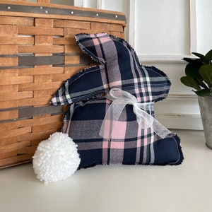May include: A blue and pink plaid bunny rabbit pillow with a white ribbon. The pillow is sitting on a white surface next to a wicker basket and a white pom-pom.