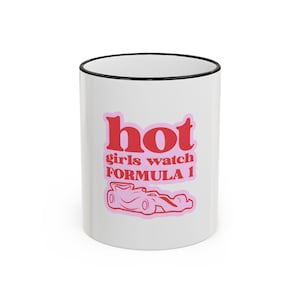 May include: White ceramic mug with a black rim, featuring the text "hot girls watch FORMULA 1" in pink lettering. A pink race car graphic is below the text. The mug is a standard size for coffee or tea.