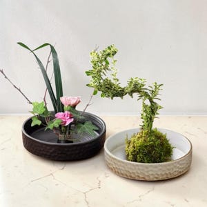 Op de afbeelding: Twee decoratieve keramische kommen met bloemstukken. Eén kom is donkerbruin met een textuur, met roze bloemen en groen. De andere is lichtbeige, met een mosachtige basis met een groen-witte plant.