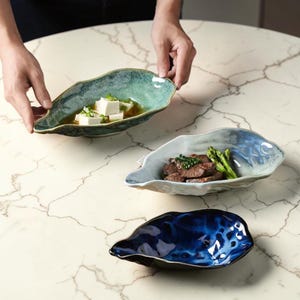 Può includere: Tre piatti da portata in ceramica a forma di foglia, nei toni del blu e del verde. Un piatto contiene tofu in brodo, un altro carne e asparagi, e il terzo è vuoto. I piatti sono su una superficie di marmo.
