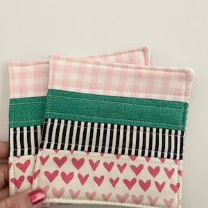May include: Two pink and green fabric coasters with a black and white stripe pattern and a pink heart pattern. The coasters are stitched together.