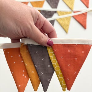 May include: A colorful banner with triangular flags in shades of orange, yellow, and brown. The flags are patterned with white crosses and small white dots. The banner is held by a hand against a white background.