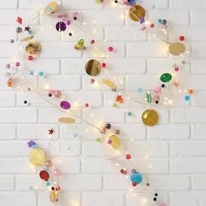May include: A decorative string of lights with colorful beads, sequins, and star-shaped accents. The garland features gold, red, purple, green, and blue elements, creating a festive and eye-catching display. The lights are strung on a thin wire.