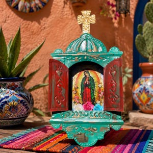 May include: A teal painted wooden retablo shrine with two hinged red doors, featuring a central image of the Virgin of Guadalupe. The shrine is topped with a gold-colored metal cross and contains three small artificial flowers at the base.