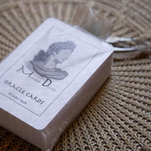 May include: A deck of "Modern Delphi Oracle Cards" wrapped in clear cellophane. The card deck has a white cover with a black and white illustration of a woman's profile and the text "Oracle Cards 75 Card Deck". The deck is tied with a ribbon.