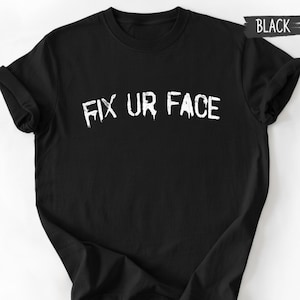 Pode incluir: Camiseta preta com o texto branco "FIX UR FACE" em uma fonte gotejante. A camiseta tem mangas curtas e gola redonda. A palavra "BLACK" está escrita em branco em uma faixa preta no canto superior direito.