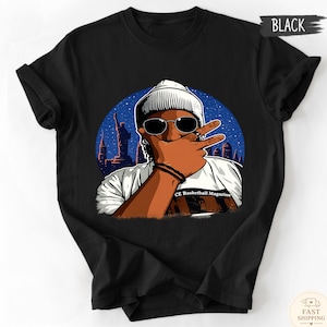 May include: A black t-shirt featuring a graphic illustration of a person wearing a beanie and sunglasses, making a peace sign gesture. The background shows a blue starry night sky with a city skyline and the Statue of Liberty. Text reads CE Basketball Magazine.