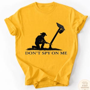 Puede incluir: Una camiseta de color amarillo mostaza con un gráfico negro de una persona con sombrero arrodillada y usando una sierra en un poste de cámara de seguridad solar. El texto DON'T SPY ON ME está impreso en letras mayúsculas negras debajo.