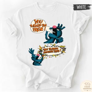 May include: White t-shirt featuring a blue monster character with a pink nose and the text "You Turned the Page!" in a speech bubble. Below, the text "You Turned Another Page!" is displayed.