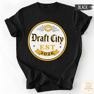 Könnte beinhalten: Schwarzes T-Shirt mit einem kreisförmigen Grafikdesign. Das Design zeigt den Text "Draft City EST 2026" in Gelb und Weiß, mit einem Football und Sternen. Das T-Shirt besteht aus einem weichen Material.