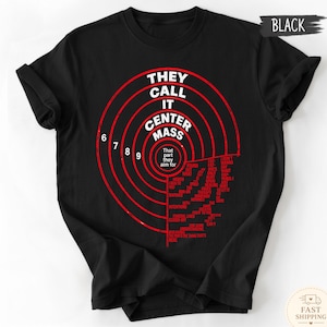 Puede incluir: Una camiseta negra de manga corta con un gráfico rojo de una diana circular. El texto dice: THEY CALL IT CENTER MASS, That part they aim for, e incluye varias frases más pequeñas dispuestas dentro de los anillos de la diana.