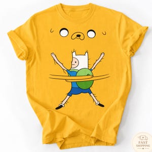 May include: Yellow t-shirt with a cartoon graphic of Finn from Adventure Time. The graphic shows Finn with his arms outstretched, wrapped in a yellow form that resembles Jake the Dog. The shirt has a crew neck and short sleeves.