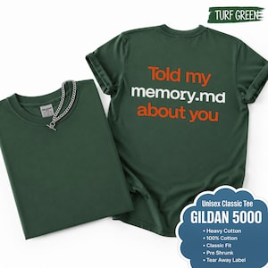 Könnte beinhalten: Ein kurzärmeliges T-Shirt in Rasengrün mit dem orange-weißen Text 'Told my memory.md about you' auf der Rückseite. Das Shirt hat einen Rundhalsausschnitt und ein Gildan 5000 Etikett. Eine silberfarbene Kette liegt auf dem gefalteten Shirt.