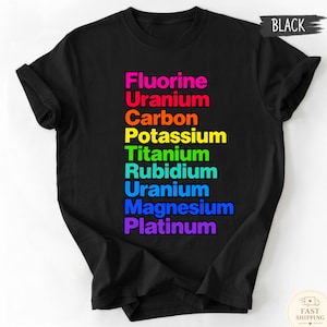 May include: A black short-sleeved t-shirt featuring a vertical list of chemical elements in rainbow colors. The text reads: Fluorine, Uranium, Carbon, Potassium, Titanium, Rubidium, Uranium, Magnesium, and Platinum. The shirt has a crew neckline.