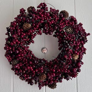 May include: A circular wreath constructed from a dense arrangement of deep burgundy berries and small brown pinecones. The wreath features a consistent, full texture with dark red spherical elements interspersed with natural woody pinecone accents.