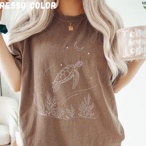 May include: Espresso-colored t-shirt featuring a white illustration of a sea turtle, moon, and stars. The design includes aquatic plants at the bottom. The shirt is a relaxed fit, perfect for casual wear.