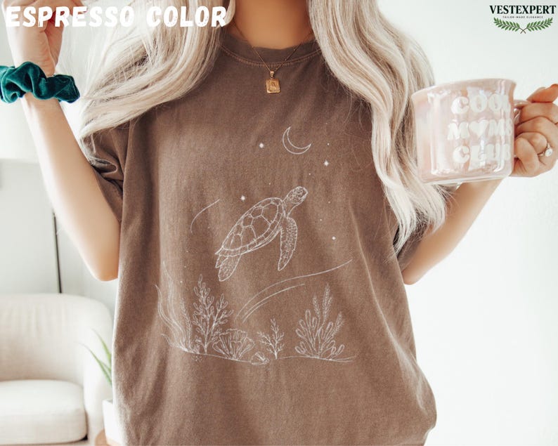 May include: Espresso-colored t-shirt featuring a white illustration of a sea turtle, moon, and stars. The design includes aquatic plants at the bottom. The shirt is a relaxed fit, perfect for casual wear.