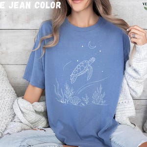 May include: Blue jean colored t-shirt featuring a white line drawing of a sea turtle swimming near coral and a crescent moon. The text "BLUE JEAN COLOR" is at the top. The shirt is a relaxed fit.