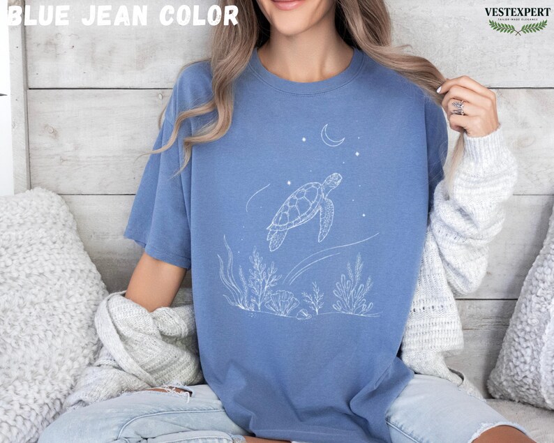 May include: Blue jean colored t-shirt featuring a white line drawing of a sea turtle swimming near coral and a crescent moon. The text "BLUE JEAN COLOR" is at the top. The shirt is a relaxed fit.
