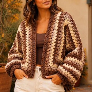 Puede incluir: Un cárdigan de ganchillo marrón, crema y beige con un ajuste relajado. El suéter abierto presenta un patrón de chevrón y mangas largas. La modelo lleva un top sin mangas marrón y pantalones color crema.
